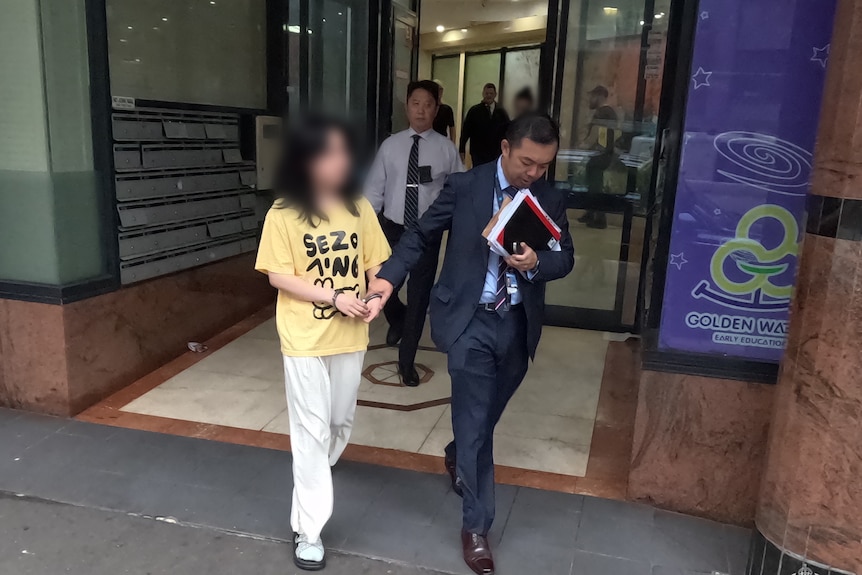 A young Chinese woman is led away in handcuffs by a detective in a blue suit. 