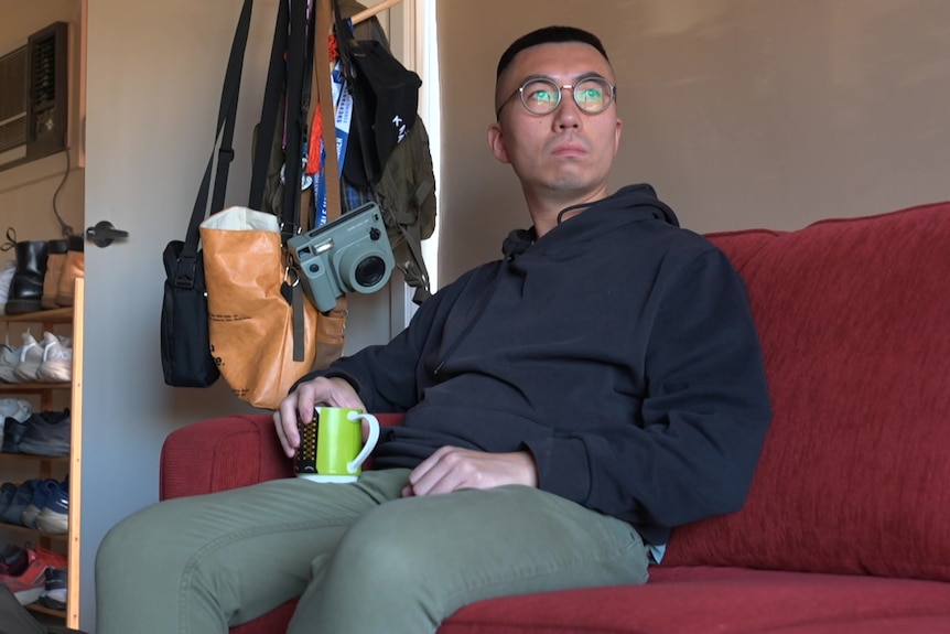A man with glasses and a dark hoodie sits and drink tea on a red sofa at home. 