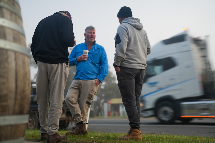 Three men drink coffee on road side grass as a truck passes behind them