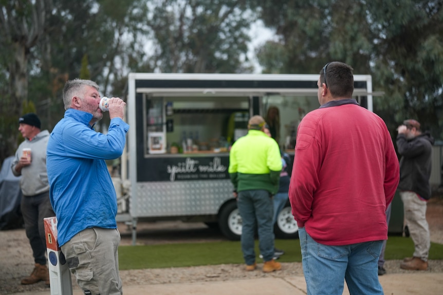 Five men standing around with coffees or waiting for coffees in front of a metal coffee van, among gum trees