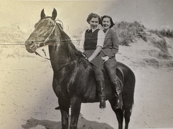 Two women ride a black horse on the beach in a black and white photo