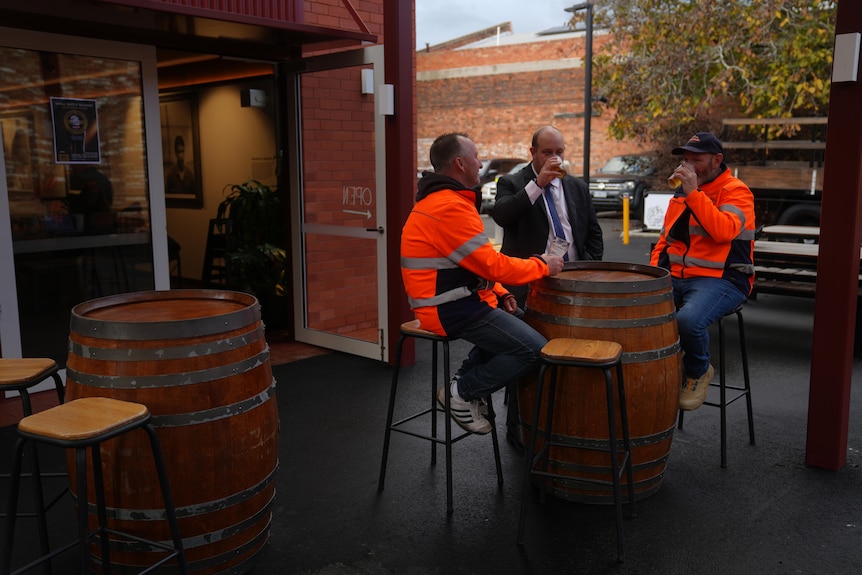 Three men sit at a barrel table and drink a beer