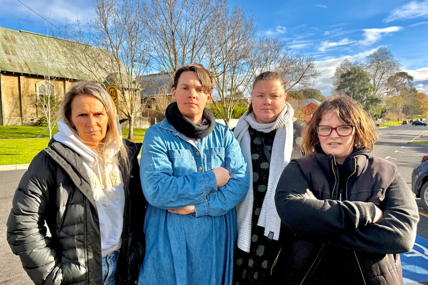 Image of a group of women with arms crossed, standing outside along a main street. 