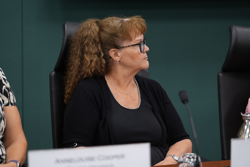A woman with curly hair, wearing glasses while sitting before a microphone.
