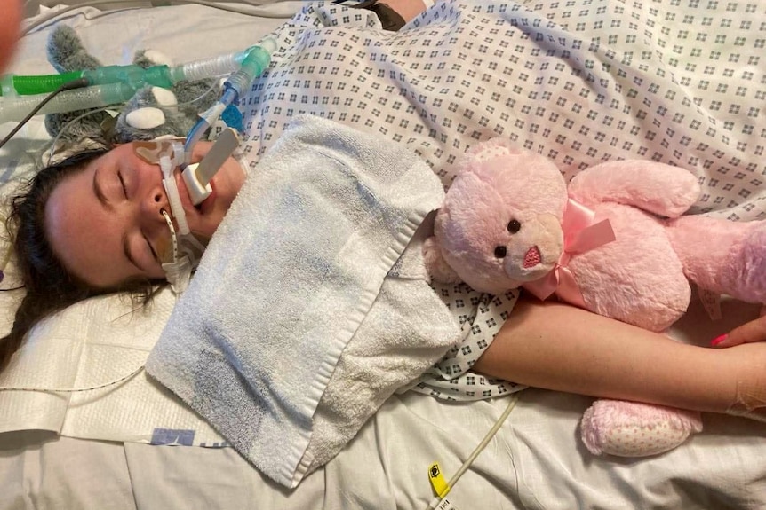 A young girl, with her eyes closed and multiple medical devices around her, in bed with her arm around a teddy bear.