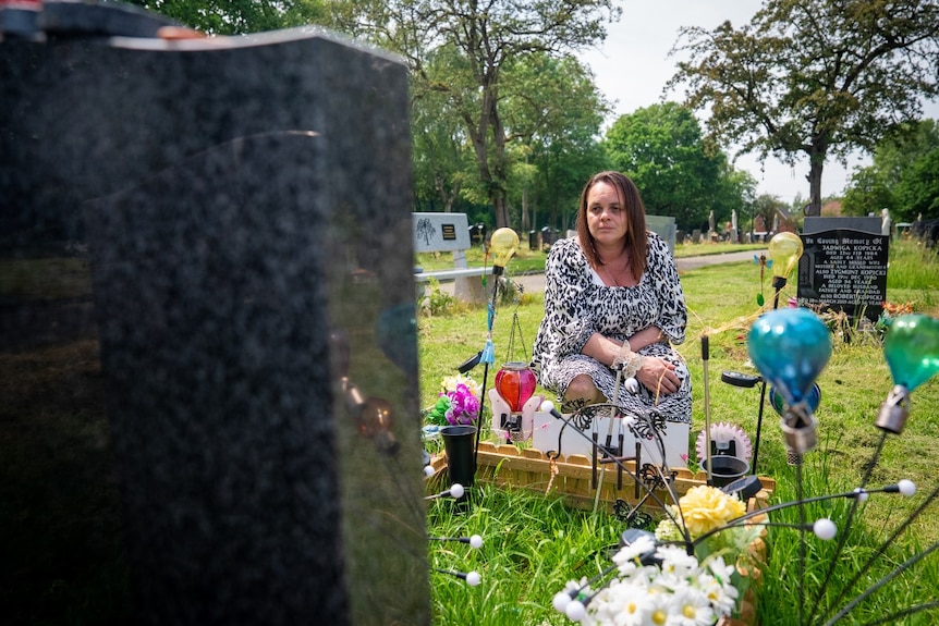 A woman kneeling and looking at a grave. She is seen from behind the grave stone.