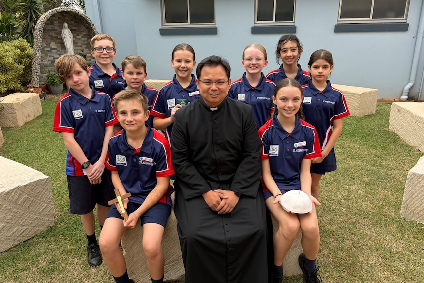 A man in a black robe sits surrounded with primary school children in a small grass courtyard