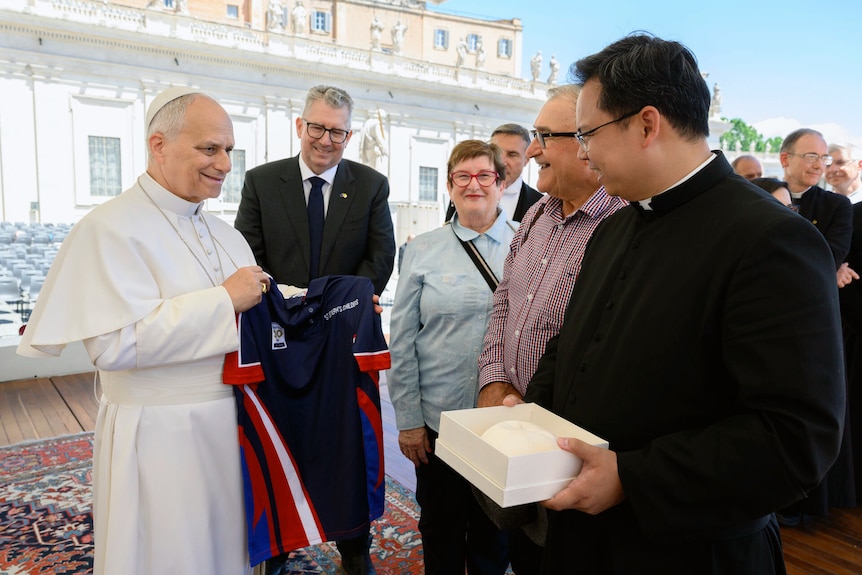 Three men and a woman stand looking at the pope dressed in white robes holding a school shirt