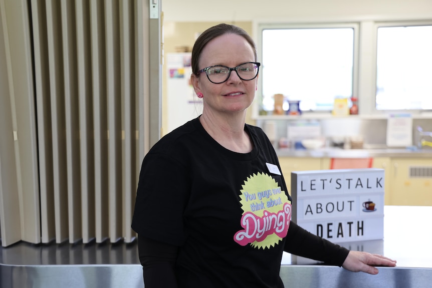 A woman in a dark t-shirt and glasses smiling at the camera with a sign reading "let's talk about death" in the background.