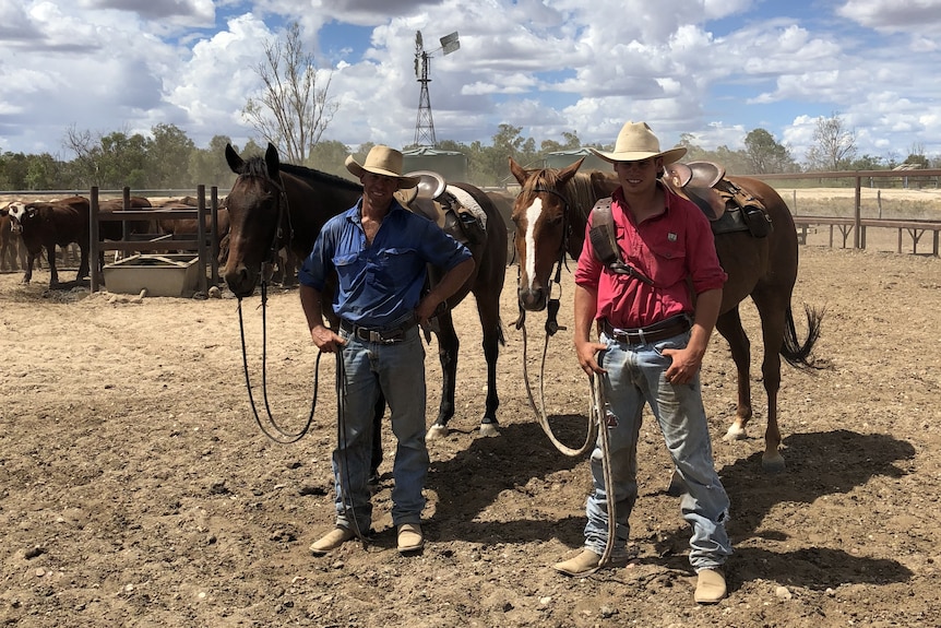 Two men, wearing jeans, shirts and Akubra-style hats, stand in a paddock holding two horses.