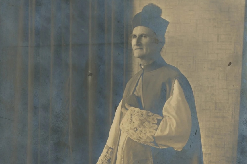 John Hawes wears a long-sleeved priest outfit, holding a small book and looks off to the distance.