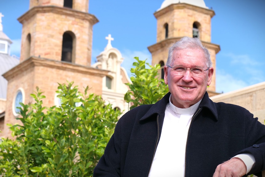 Father Cross smiles as he stands in front of plants and the cathedral. He wears glasses and a collared shirt.